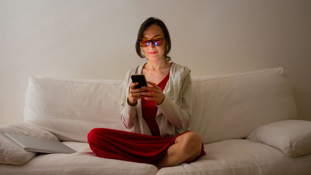 woman in a dimly light room wearing blue light blocking glasses as she reads her phone