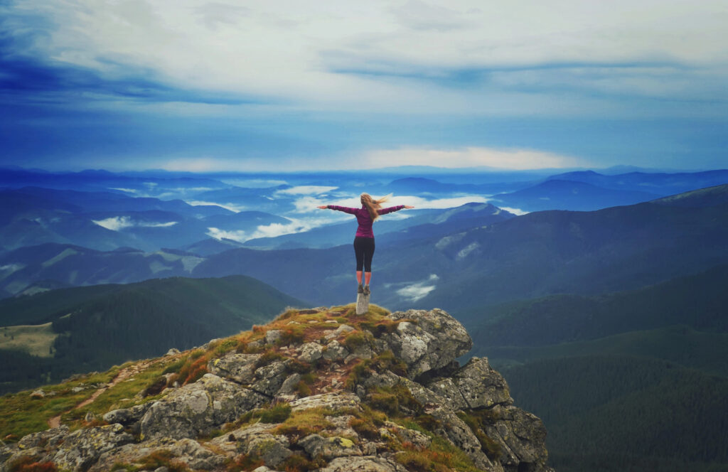 A woman standing on a mountain top with her arms wide open and the scenes of mountain peaks are in front of her